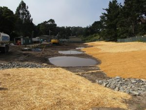 Construction site with straw-covered ground, puddles, machinery, and trees in the background.