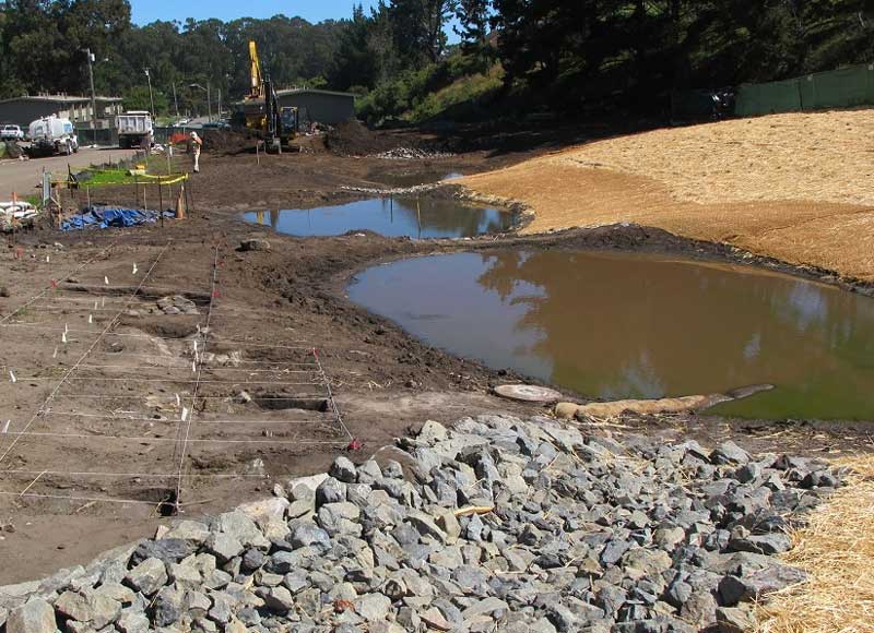 A construction site with a pond, rocks in the foreground, and straw-covered ground on the right.