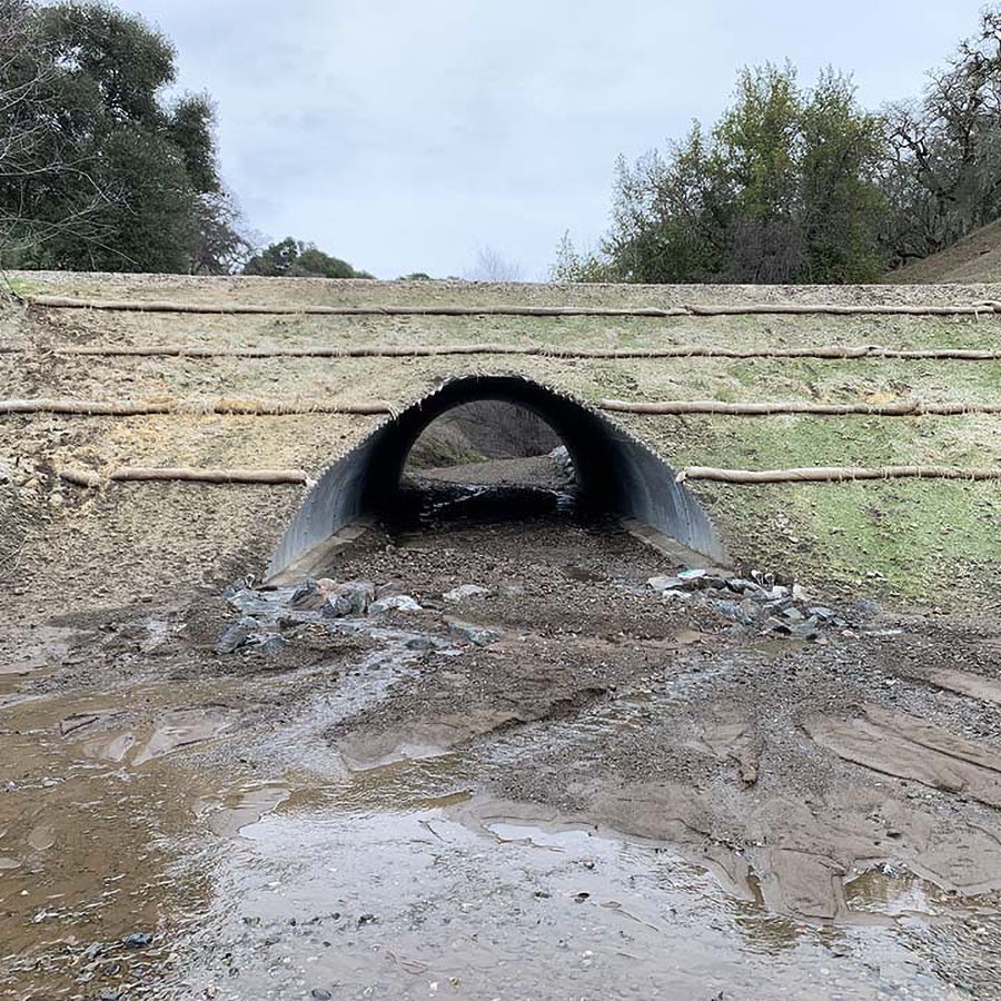 A large drainage pipe runs under terraced earth with water flowing out and small rocks scattered below.