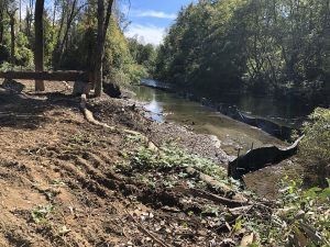 Dirt path and wooden bench beside a river, with trees and erosion control barriers along the shoreline.