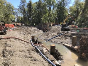 Construction site by a creek with machinery, workers, and pipes amid trees and dirt mounds.
