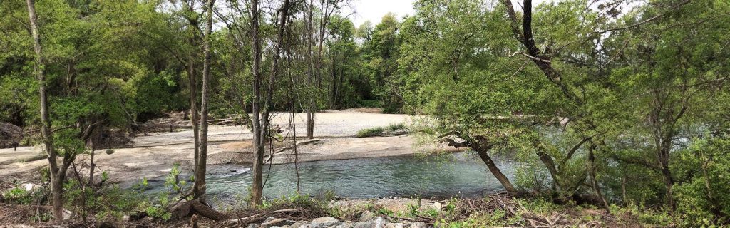 A river flows through a forest with green trees and a sandy, open embankment on a cloudy day.