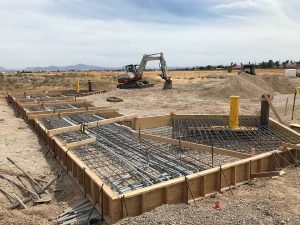 Construction site with rebar and wooden forms, excavator and dirt piles in the background, under a cloudy sky.