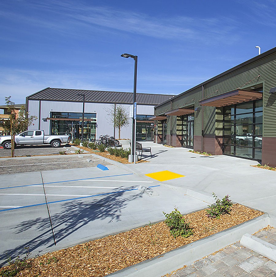 Modern commercial buildings with large windows, parking spaces, and a white pickup truck under a clear blue sky.