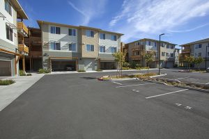 Modern apartment buildings with garages and a mostly empty parking lot under a bright blue sky.