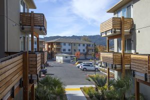 Modern apartment complex with wooden balconies, parked cars, and mountains in the background under a blue sky.
