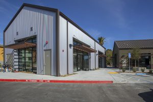 Modern gray metal building with large windows, next to a parking lot and a marked fire lane curb.