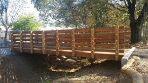 A wooden footbridge with sturdy railings spans over a shallow, dry creek bed in a wooded area.