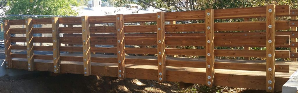 A wooden pedestrian bridge with sturdy railings and metal bolts, surrounded by trees and sunlight.