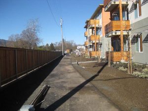 A paved path runs beside a wooden fence and modern apartment buildings on a clear, sunny day.
