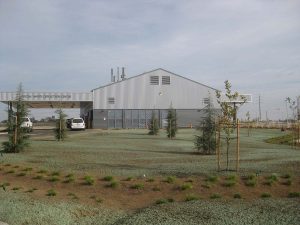 A modern industrial building with parked cars, young trees, and sparse landscaping in front.