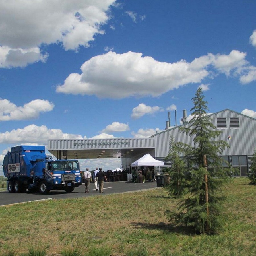 A blue garbage truck near a gray industrial building under a blue sky with clouds.