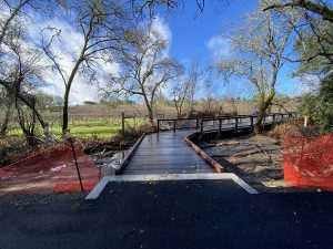 Wooden footbridge with red safety fencing on both sides, leading into a scenic, tree-lined rural area.