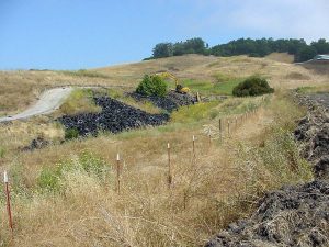 Excavator working near a large pile of tires in a grassy, hilly landscape with scattered trees.