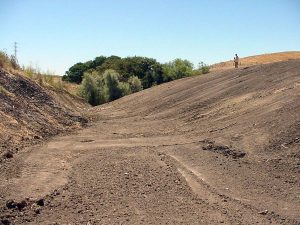 A dry, barren landscape with a person standing on a sloped dirt embankment under a clear sky.