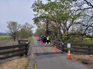 Construction workers repair a wooden bridge on a rural trail, with orange cones blocking the path.
