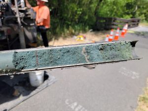 Close-up of a soil core sample with greenish layers, blurred worker and traffic cones in background.