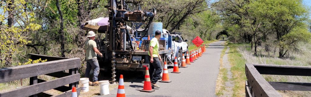 Two workers operate machinery on a trail, surrounded by traffic cones and a truck, with trees in the background.