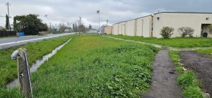 A grassy area with a narrow ditch beside a road and a beige building; an American flag in the distance.
