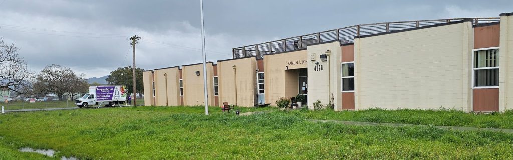 Single-story beige building with a sign reading SAMUEL CUN...; truck and green grass in front, cloudy sky.