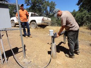 Two men work outdoors near a truck; one inspects equipment on a well pipe, the other stands watching.
