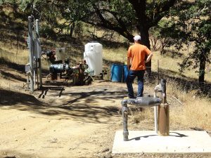 A man in an orange shirt stands near water equipment and tanks in a rural outdoor setting.