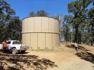 Large beige water tank near a dirt road, with a pickup truck and two people in a wooded area.