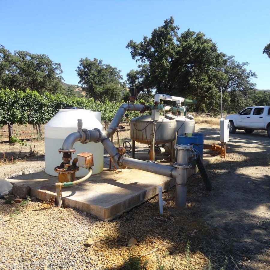 Irrigation system with pipes, tanks, and controls near a vineyard and parked white truck outdoors.