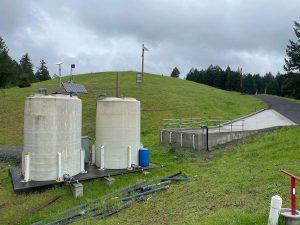 Two large water tanks and pipes sit on grass near a small concrete ramp, under a cloudy sky.
