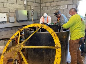 Three workers wearing safety gear inspect or repair equipment inside a large industrial tank indoors.
