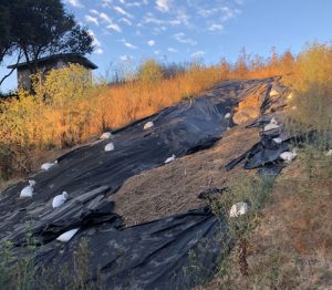 Plastic sheeting held by sandbags covers a grassy hill under a blue sky with wispy clouds.