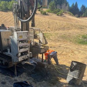 A worker in an orange vest operates drilling equipment on a dry, grassy hillside.