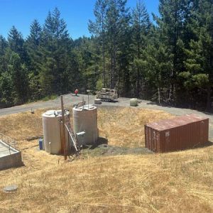Two large water tanks and a rusted shipping container sit in a dry grassy area surrounded by tall trees.