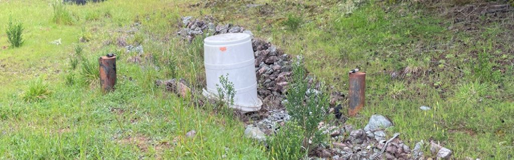 Large white barrel surrounded by rocks and grass, flanked by two rusted metal pipes on either side.