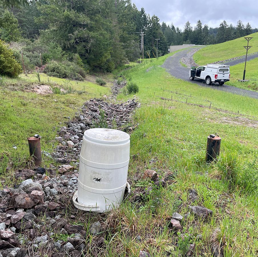 A white barrel sits among rocks in a grassy field near a parked white truck and a sloped road.