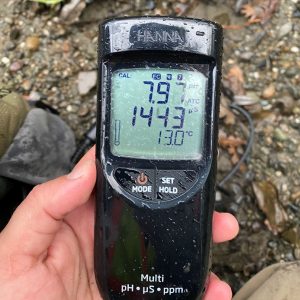 A hand holds a digital water quality meter showing pH 7.87, EC 1443 µS, and temperature 13.0°C.