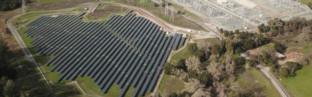 Aerial view of a large solar panel array near a power substation, surrounded by greenery and trees.
