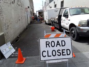 Narrow alleyway blocked by a Road Closed sign, cones, work trucks, and construction workers ahead.