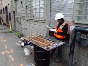 A construction worker inspects soil core samples on a table outside a building, taking notes.