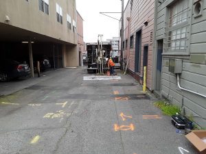 Worker operates drilling equipment in an urban alley with markings and cars parked to the side.