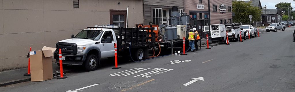 Utility workers with trucks and equipment work on a street, with traffic cones blocking off the area.
