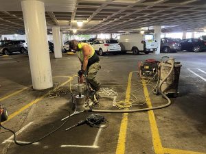 Worker in safety gear uses drilling equipment on the floor of a parking garage, surrounded by tools and cables.