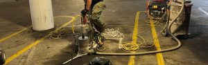 Worker operating heavy drilling equipment on a concrete floor in an industrial parking garage.