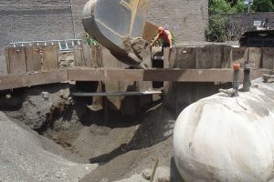 Excavator pouring sand near a large underground tank at a construction site with a worker standing nearby.