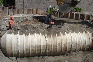 Two construction workers install and cover a large horizontal cylindrical tank in a dirt excavation site.