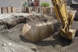Excavator lifting soil near a large, partially buried metal tank at a construction site.