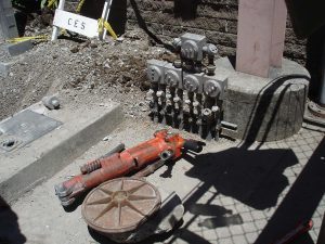 Several industrial valves and pipes next to a red jackhammer and a metal wheel on a construction site.