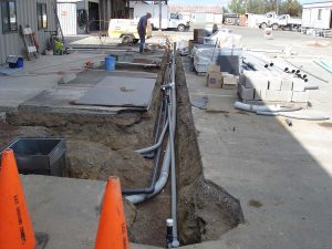 Construction site with exposed pipes in a trench, cones, materials, and a worker in the background.