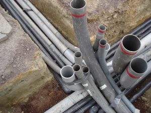 A cluster of gray PVC pipes emerges from the ground at a construction site with exposed soil and trenches.