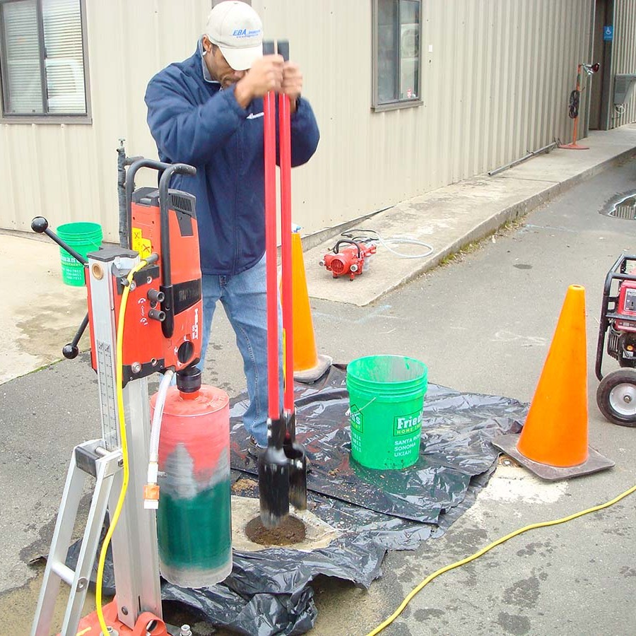 A worker uses a manual tool to dig a hole outside, surrounded by safety cones and equipment.